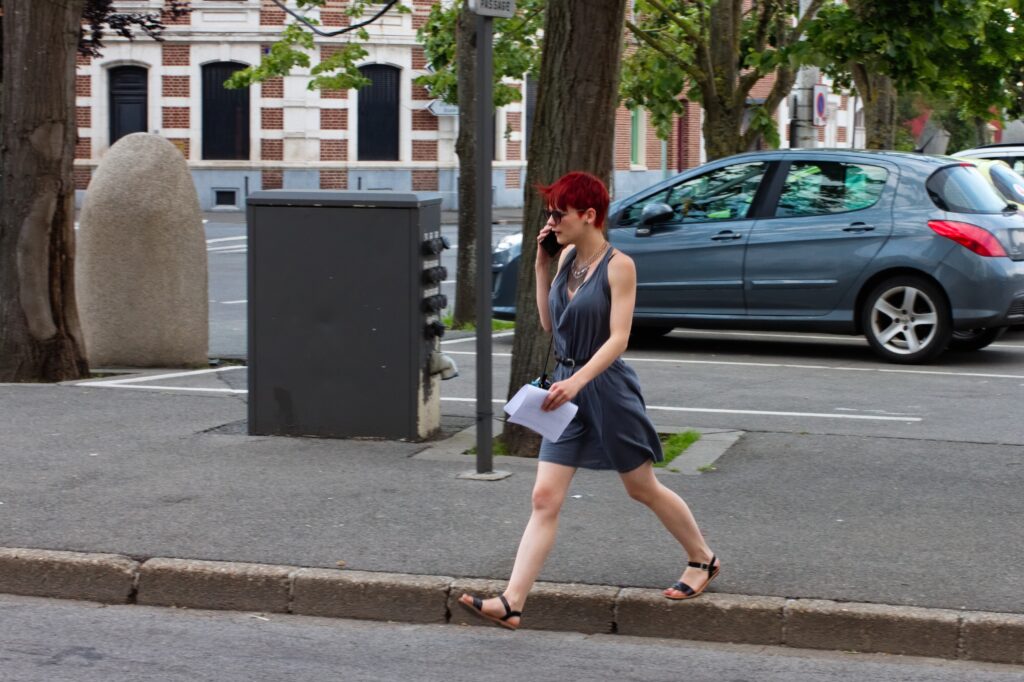Jeune femme rousse en robe qui marche d’un pas vif, des documents à la main et le téléphone à l’oreille. En arrière une place avec une voiture garée et des arbres.